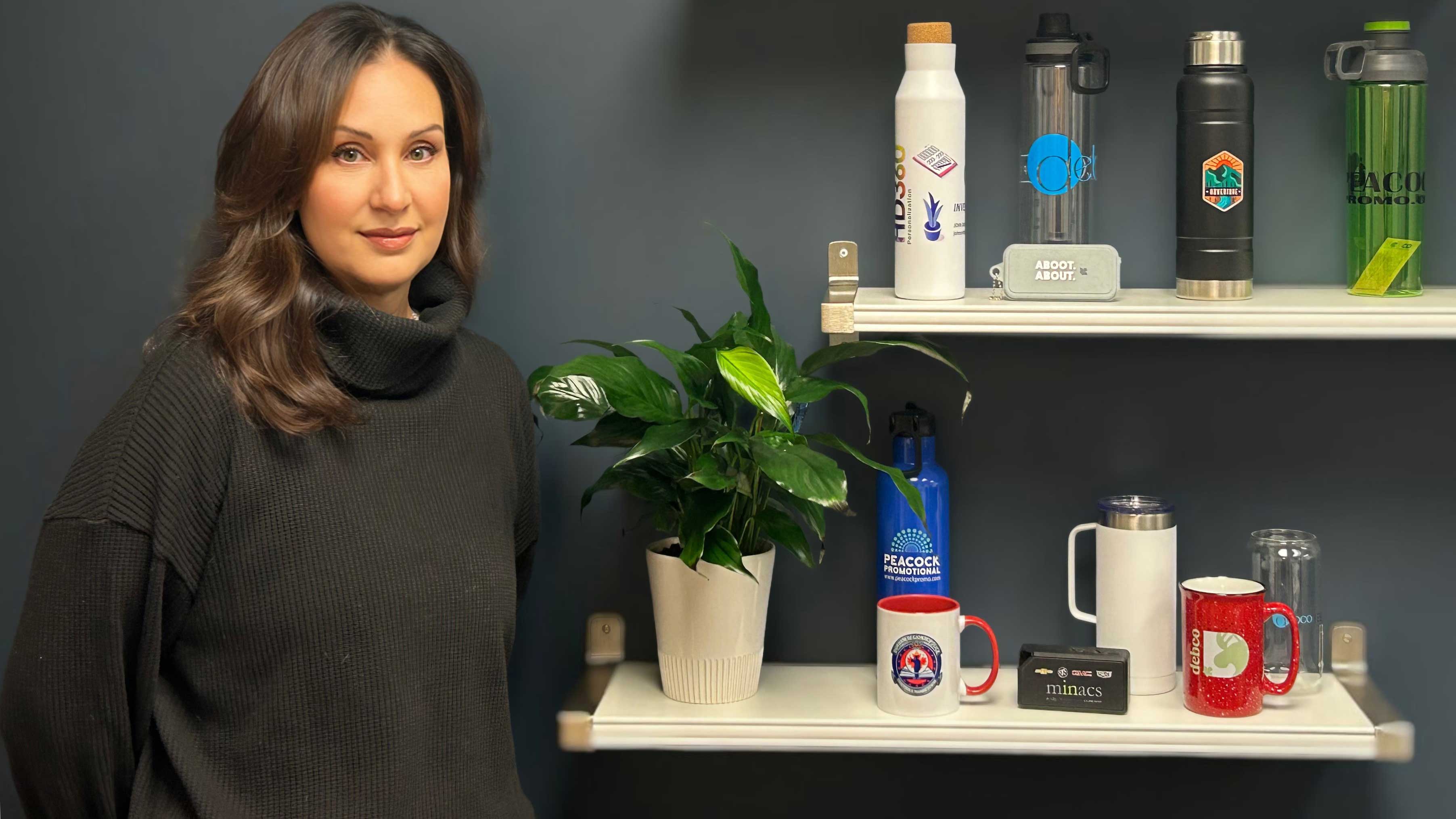 Owner Melissa Peacock, smiling and standing beside two shelves of promotional products