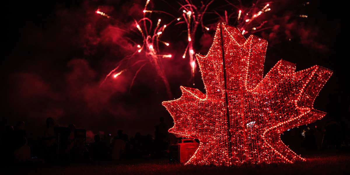 Fireworks in the sky with a lit up maple leaf in the foreground.